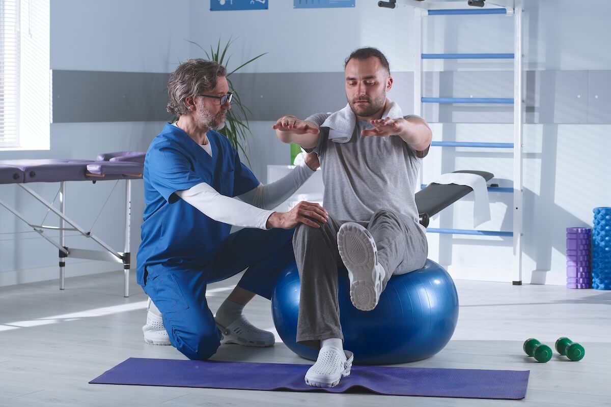 A physiotherapist assists a man as he balances on an exercise ball, promoting strength and stability in a supportive environment.